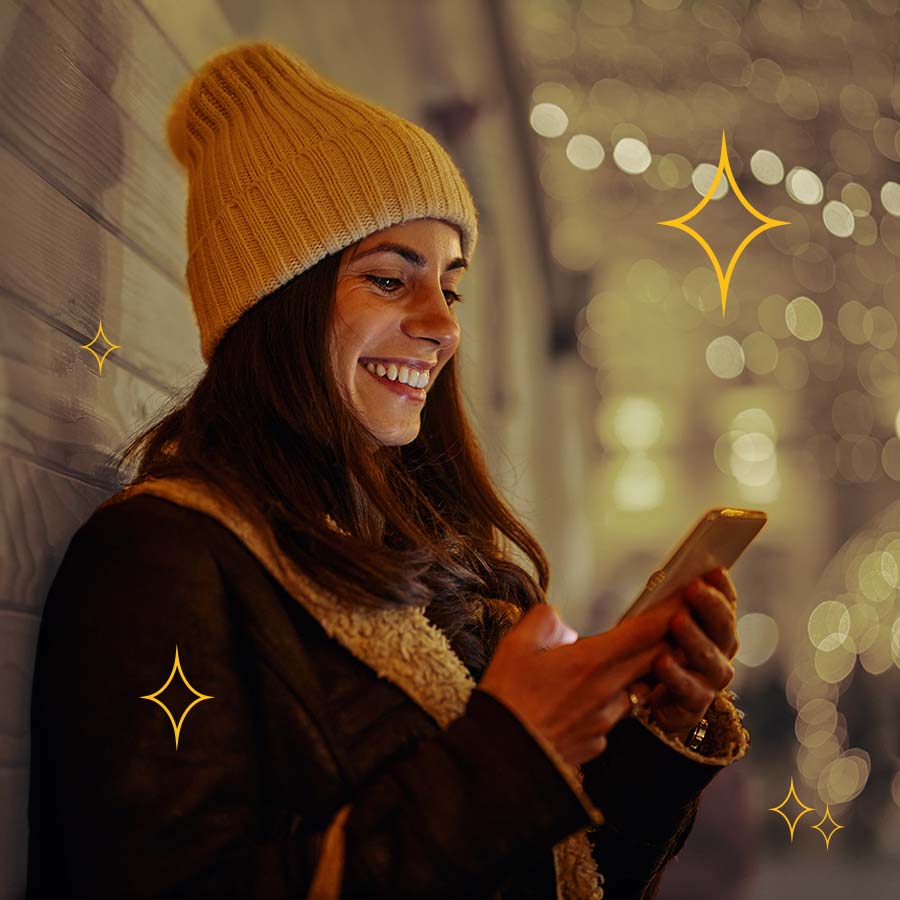 A woman in a yellow beanie smiles at her phone while leaning against a wall. The background is blurred with festive lights and silhouettes of people.
