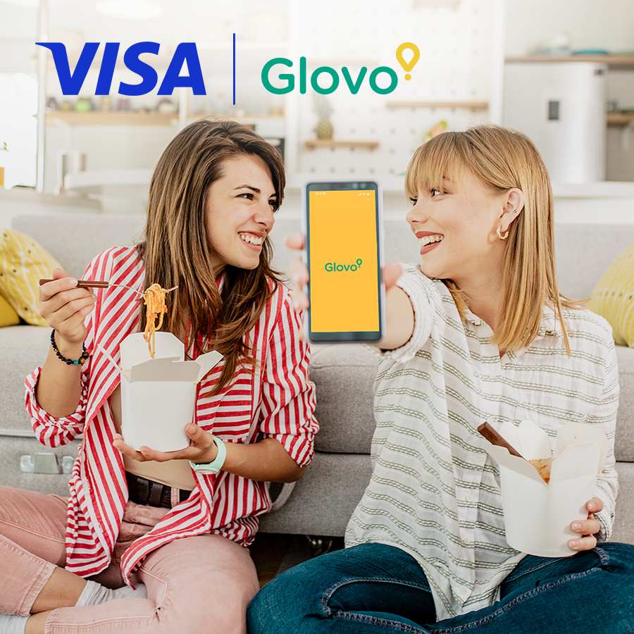 Two women smiling and sitting on a couch, one holding a smartphone with the Glovo app, the other holding noodles. Visa and Glovo logos are displayed.