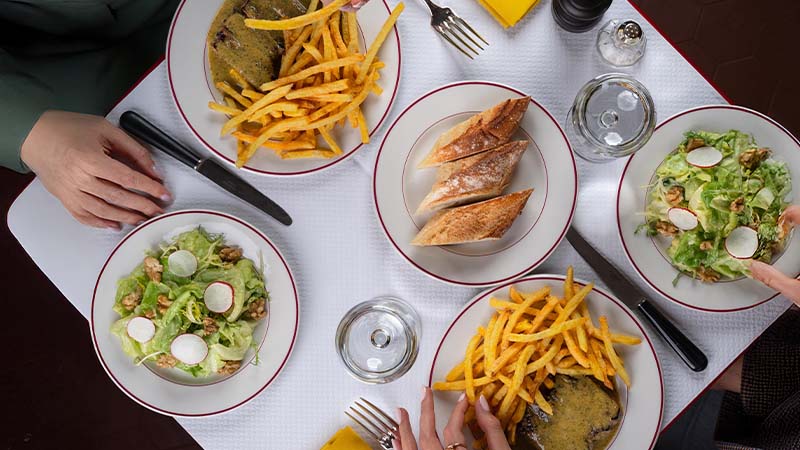 Overhead view of a table set with plates of salad, steak with fries, sliced bread, cutlery, glassware, and two diners’ hands reaching for food.