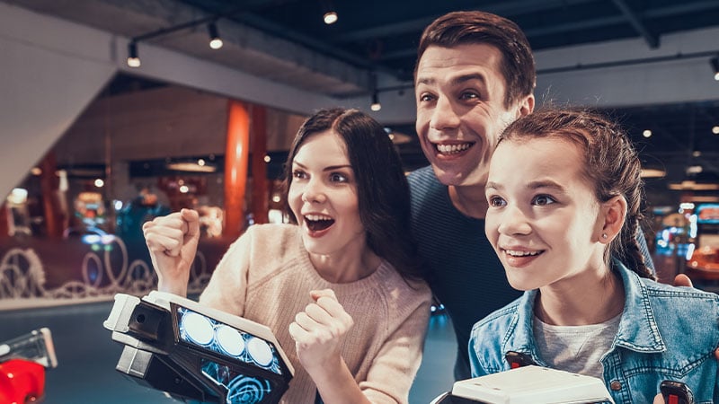 A group of three people, including two young women and a man, play arcade games together and appear excited.