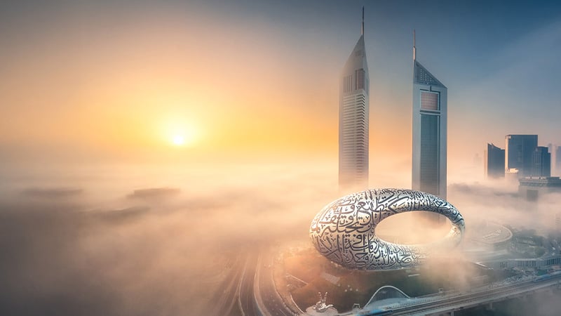A futuristic building with Arabic calligraphy, the Museum of the Future, stands amid fog near two skyscrapers in Dubai at sunrise.
