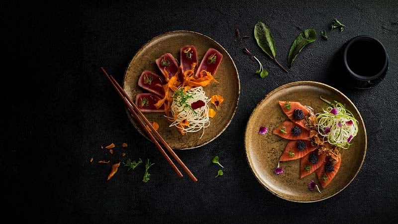Two ceramic plates with artfully arranged tuna sashimi, noodles, garnishes, and edible flowers on a dark surface; chopsticks and a small cup are also visible.