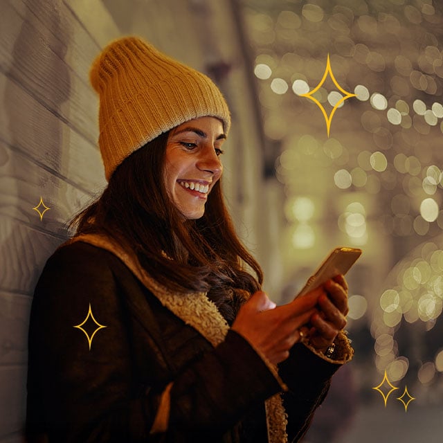 A woman in a yellow beanie smiles at her phone while leaning against a wall. The background is blurred with festive lights and silhouettes of people.
