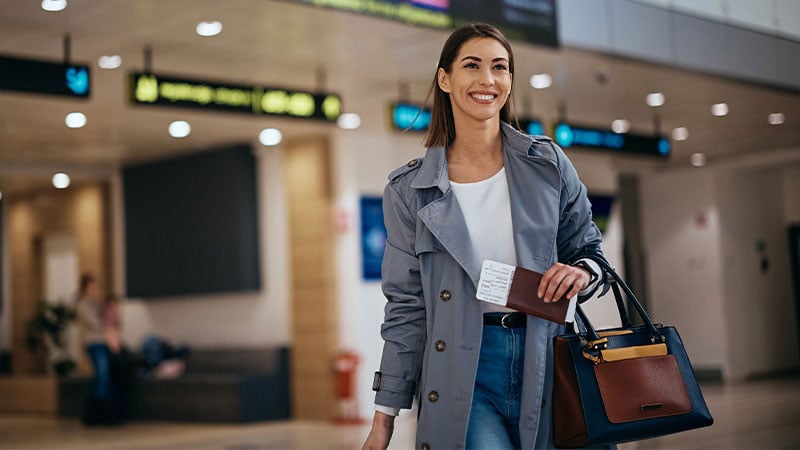 A smiling woman in a gray coat holds a passport and boarding pass in an airport. She carries a stylish handbag, conveying a sense of travel and anticipation.