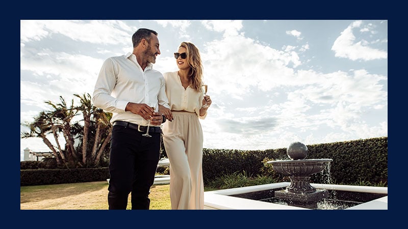A couple in elegant attire walks beside a garden fountain, holding champagne glasses. They look happy and relaxed under a partly cloudy sky.